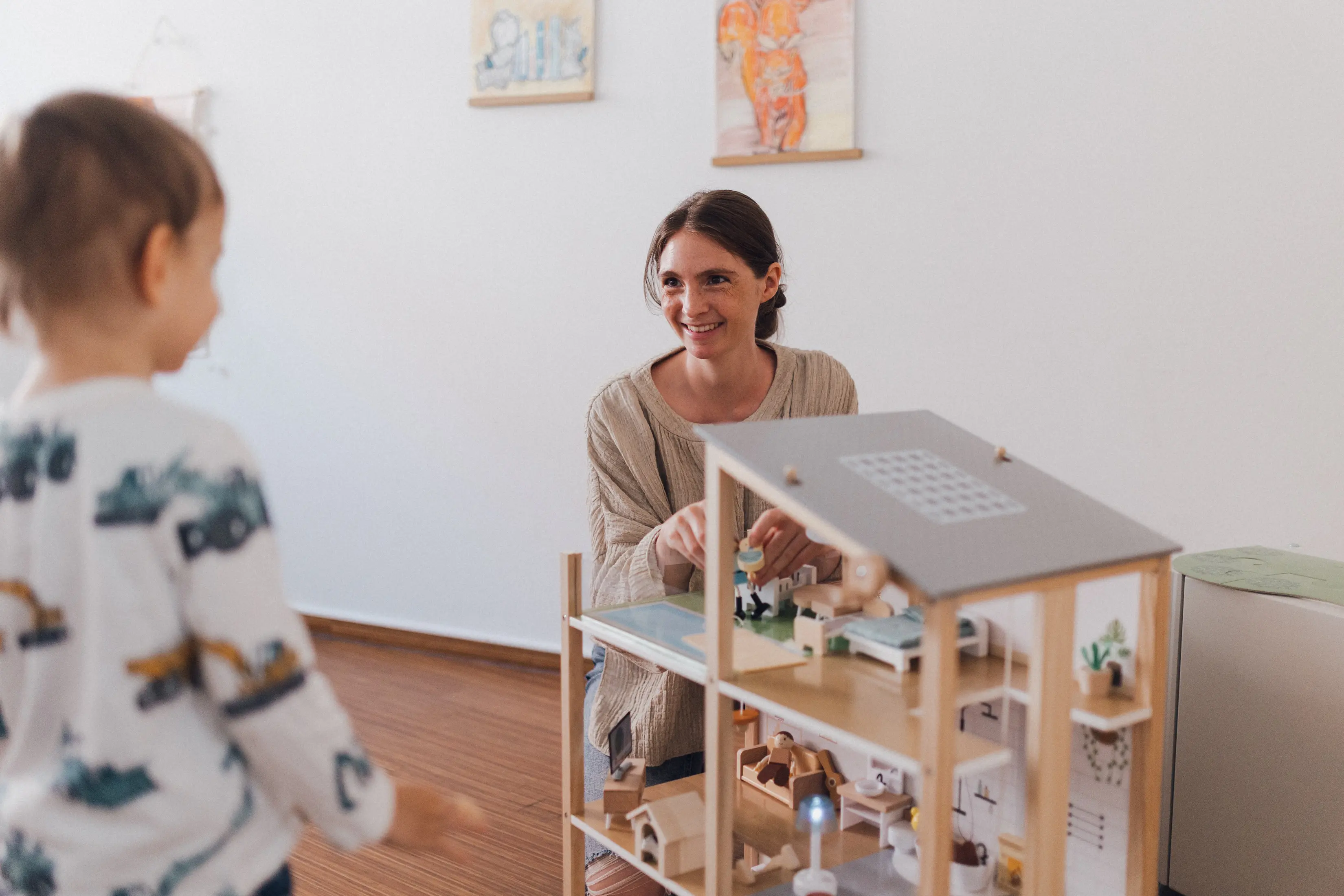 Kind steht vor einem Puppenhaus neben einer Logopädin